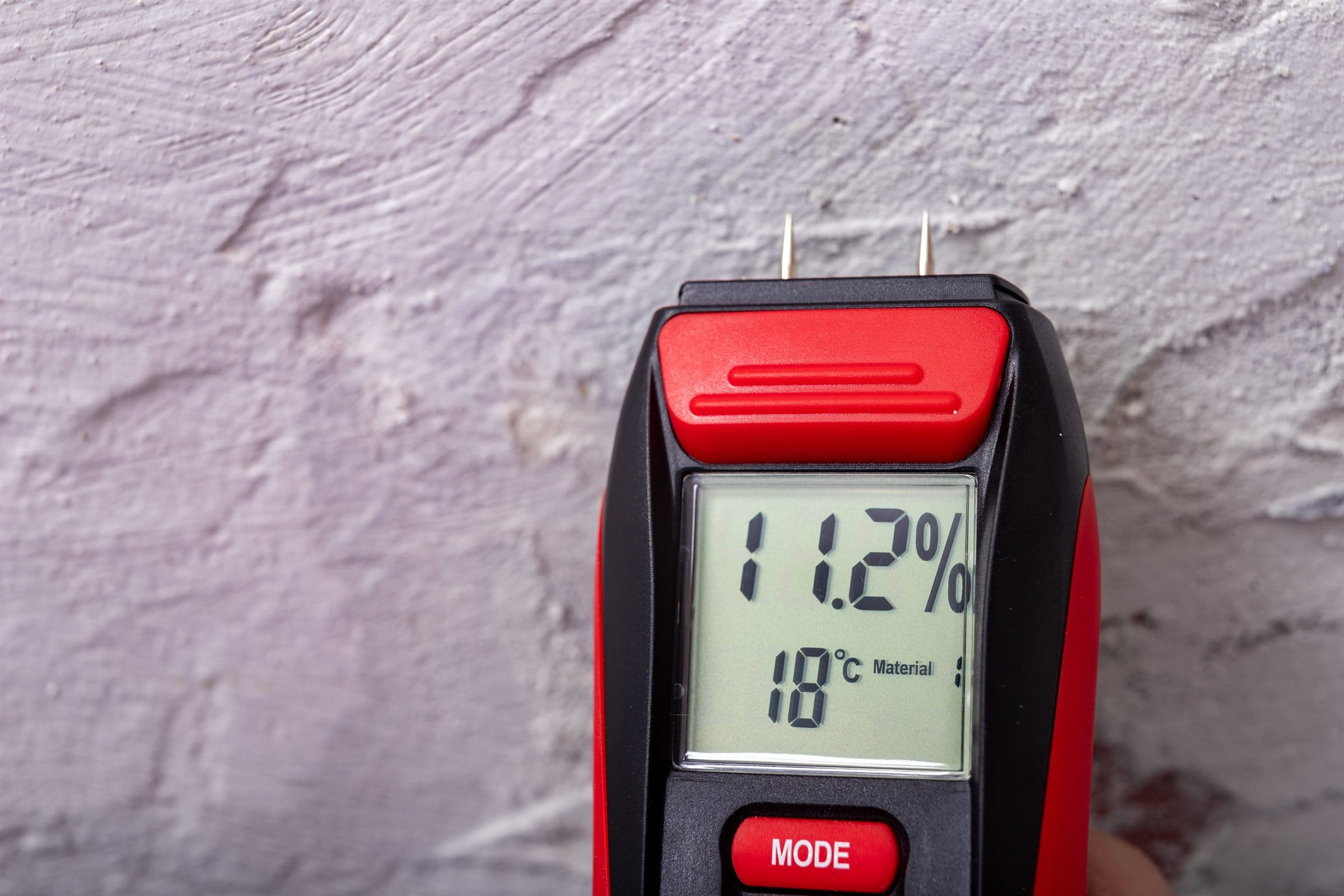 A handheld electronic moisture meter held against a plaster wall, its display reading 11.2 percent moisture content (illustrative)