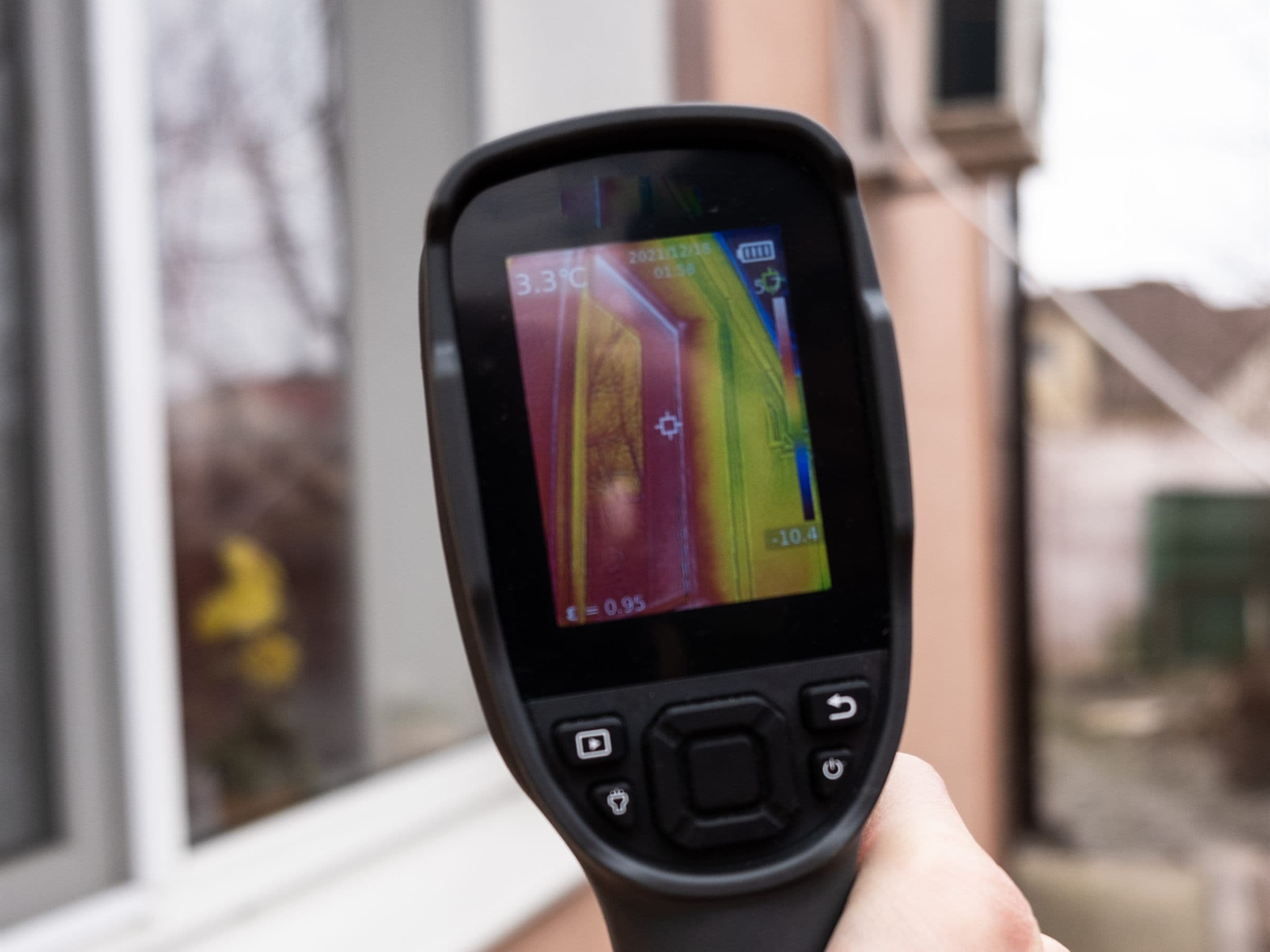 A handheld thermal imaging camera held up to a house window, a false-colour heat image of the window frame showing on its screen (illustrative)